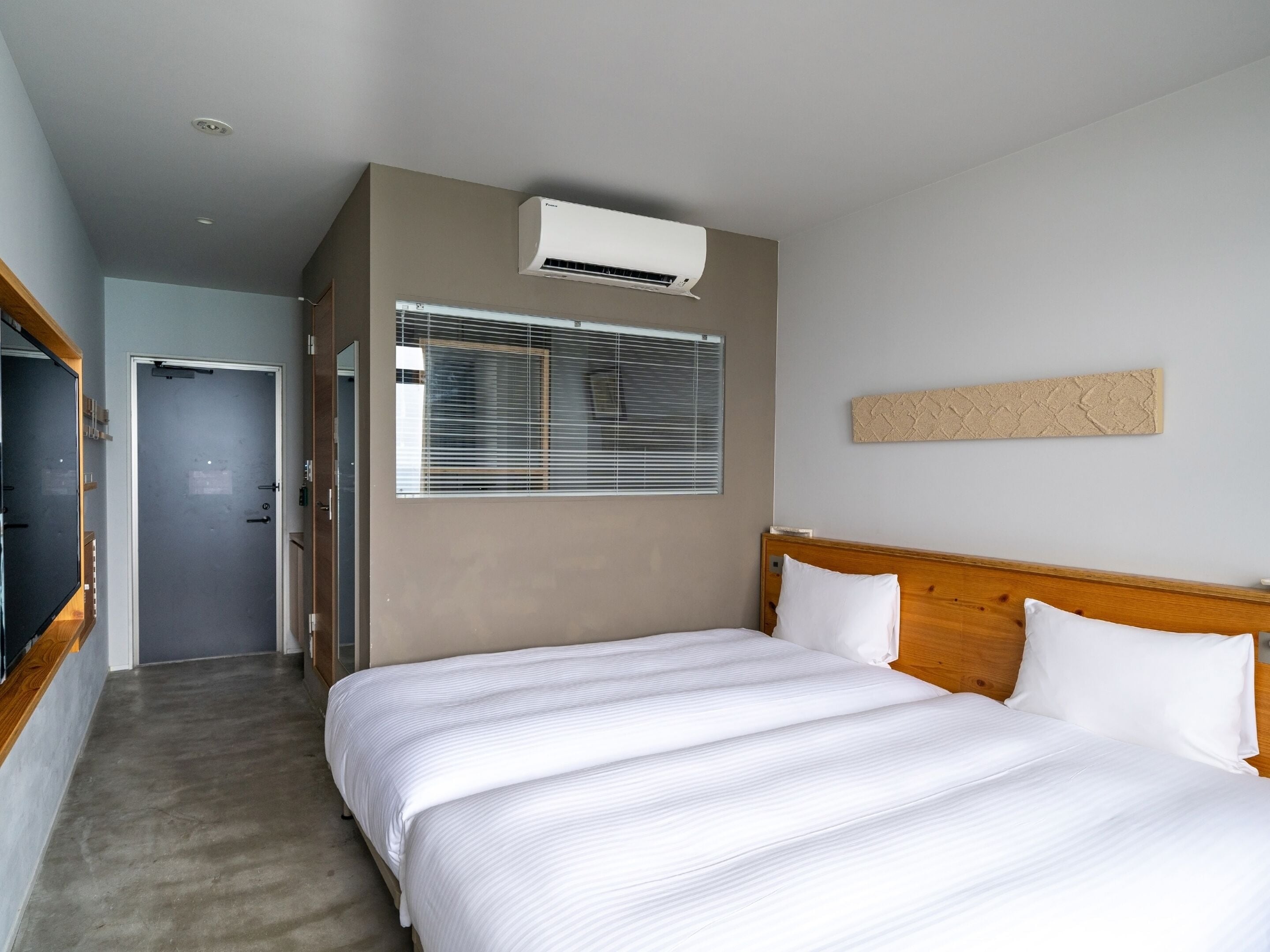 A hotel room with a bed, a window with blinds, an air conditioning unit, and a wooden headboard.