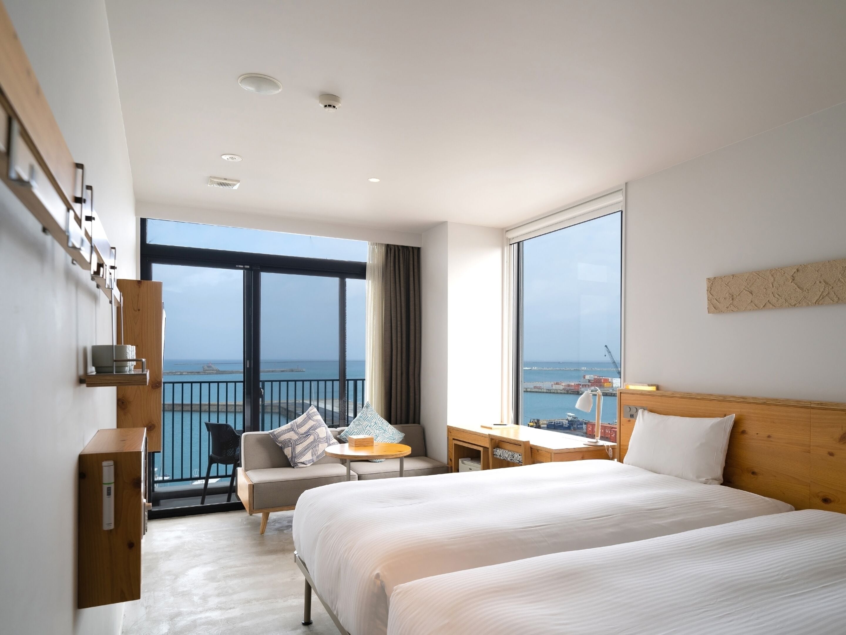 A modern hotel room with a large bed, a desk, a chair, and a view of the sea through a balcony.