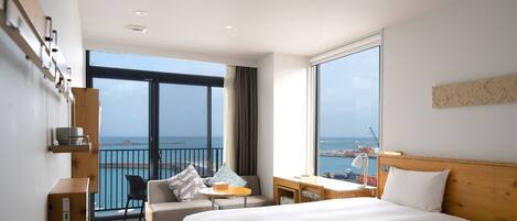 A modern hotel room with a large bed, a desk, a chair, and a view of the sea through a balcony.