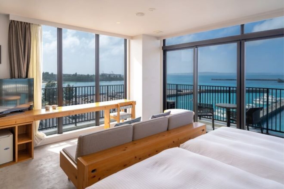 A hotel room with a large bed, a desk with a TV, and a balcony with a view of the sea.