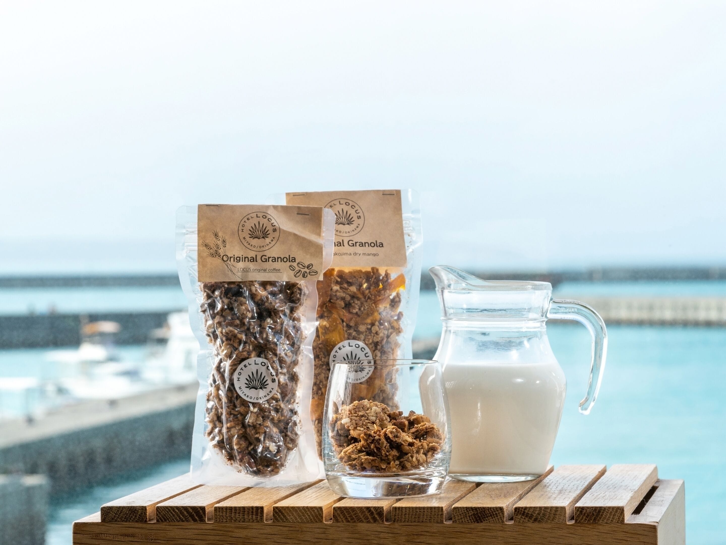 Two bags of granola and a glass pitcher of milk on a wooden table with a view of the sea.