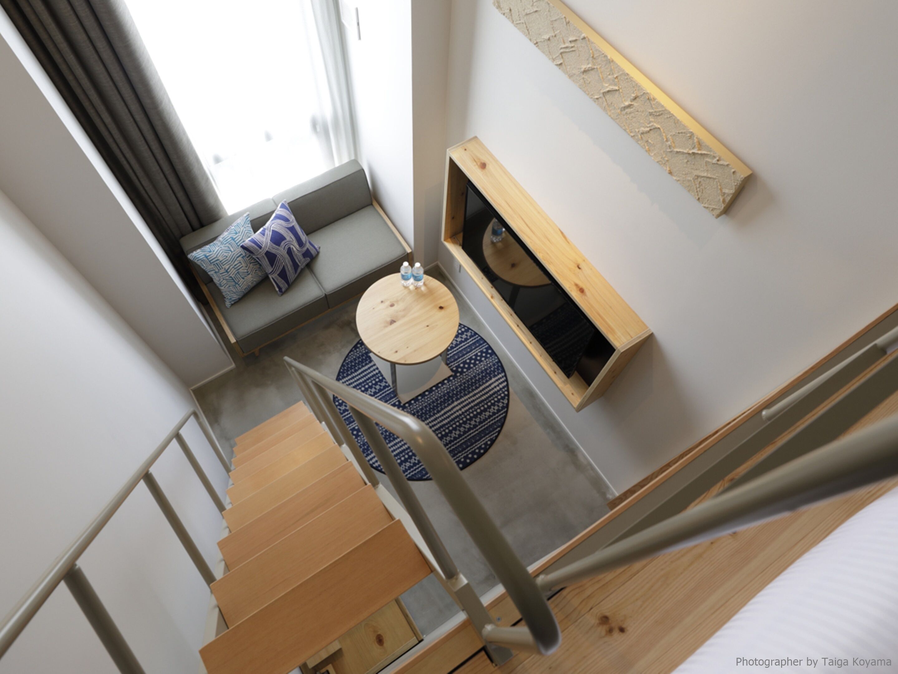A modern interior with a wooden staircase, a round wooden table, a sofa with patterned cushions, and a small window with a view.