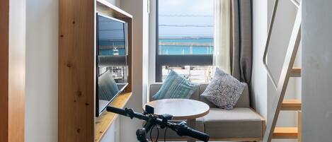 Habitación compacta con una bicicleta, un televisor fijado en un panel de madera, un sofá con cojines estampados y vistas al mar a través de amplios ventanales.
