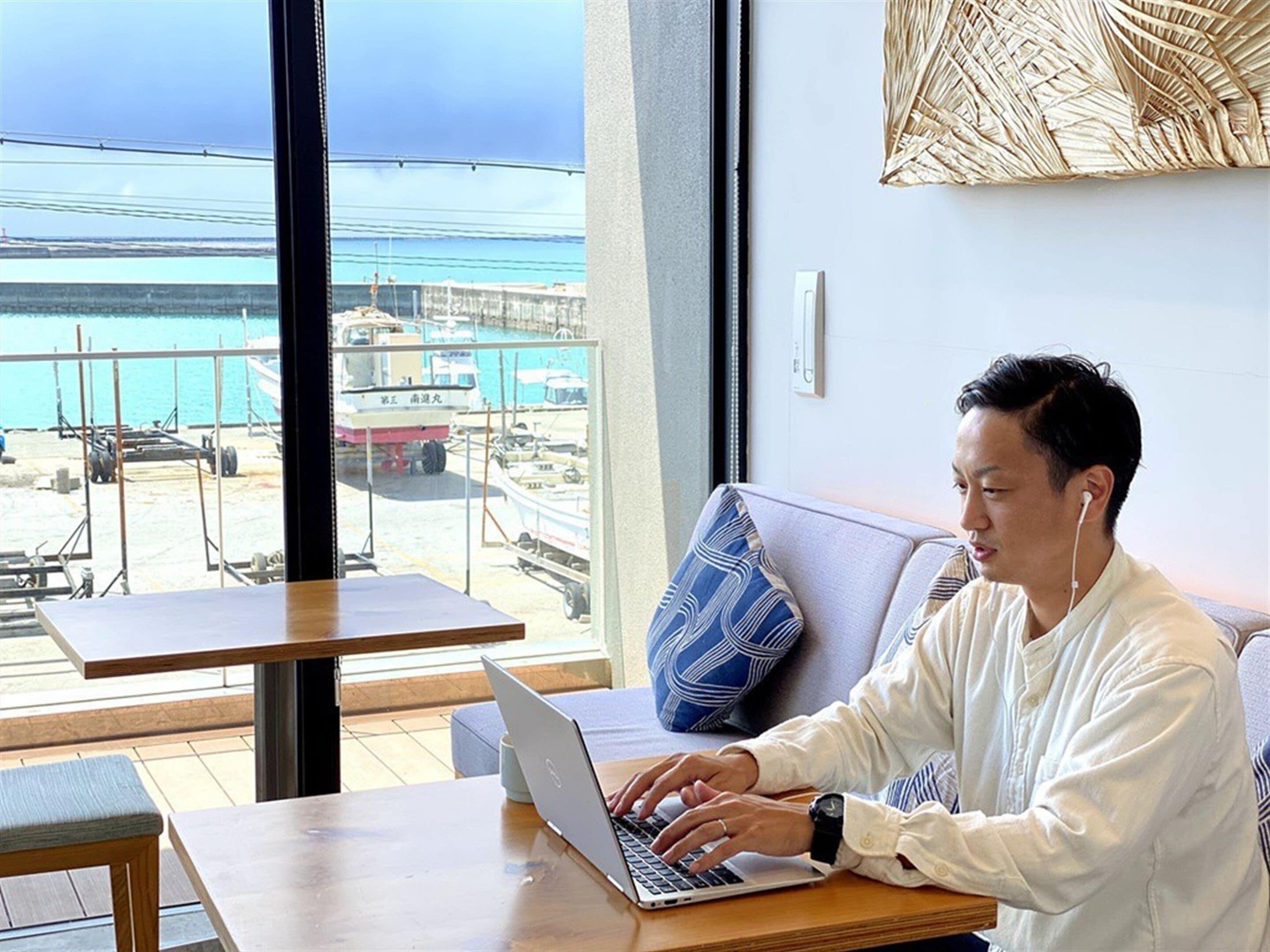 Une personne travaille sur un ordinateur portable, assise à une table en bois, avec vue sur une marina et des bateaux, le tout visible à travers une grande fenêtre.