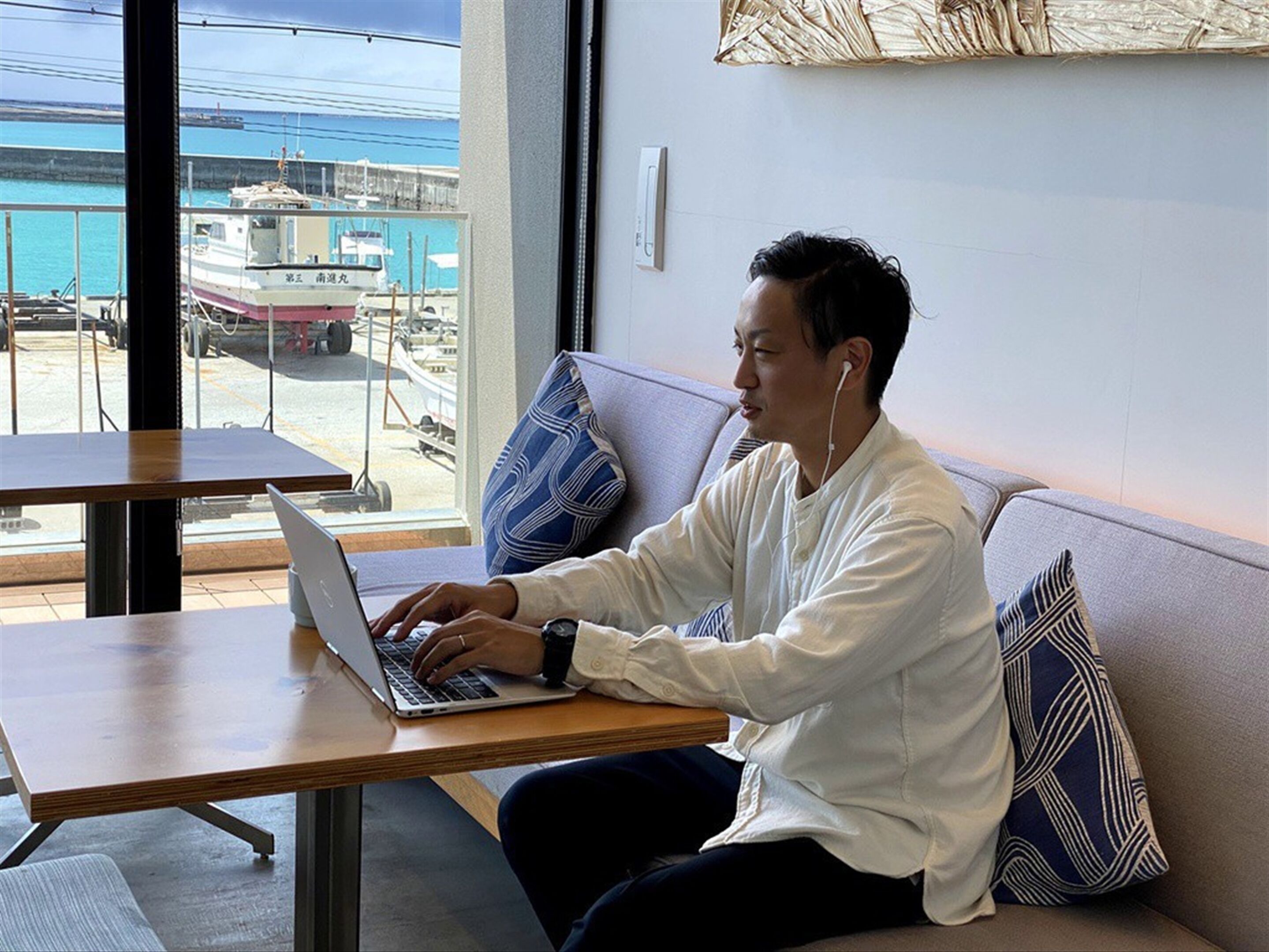 Eine Person sitzt an einem Tisch mit einem Laptop, im Hintergrund ist ein Hafen mit Booten zu sehen.