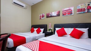 Habitación de hotel con dos camas, cada una con ropa de cama blanca y roja, y una manta estampada en blanco y rojo. Se observa un aire acondicionado instalado en la pared y tres cuadros que muestran un autobús rojo, un autobús de dos pisos rojo y una persona con una maleta.