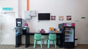 Una kitchenette con cafetera, refrigerador, dos sillas verdes y un televisor en la pared.