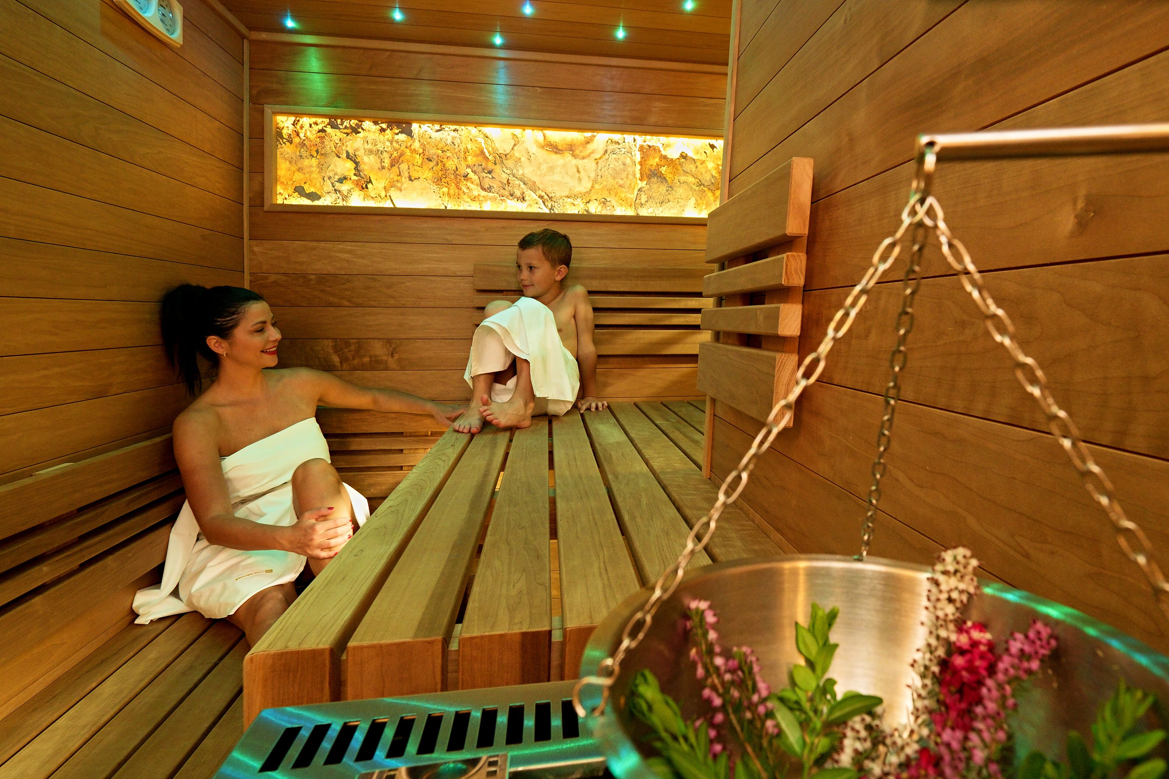 A wooden sauna with benches, a hanging metal container with flowers, and a window with a view of trees.