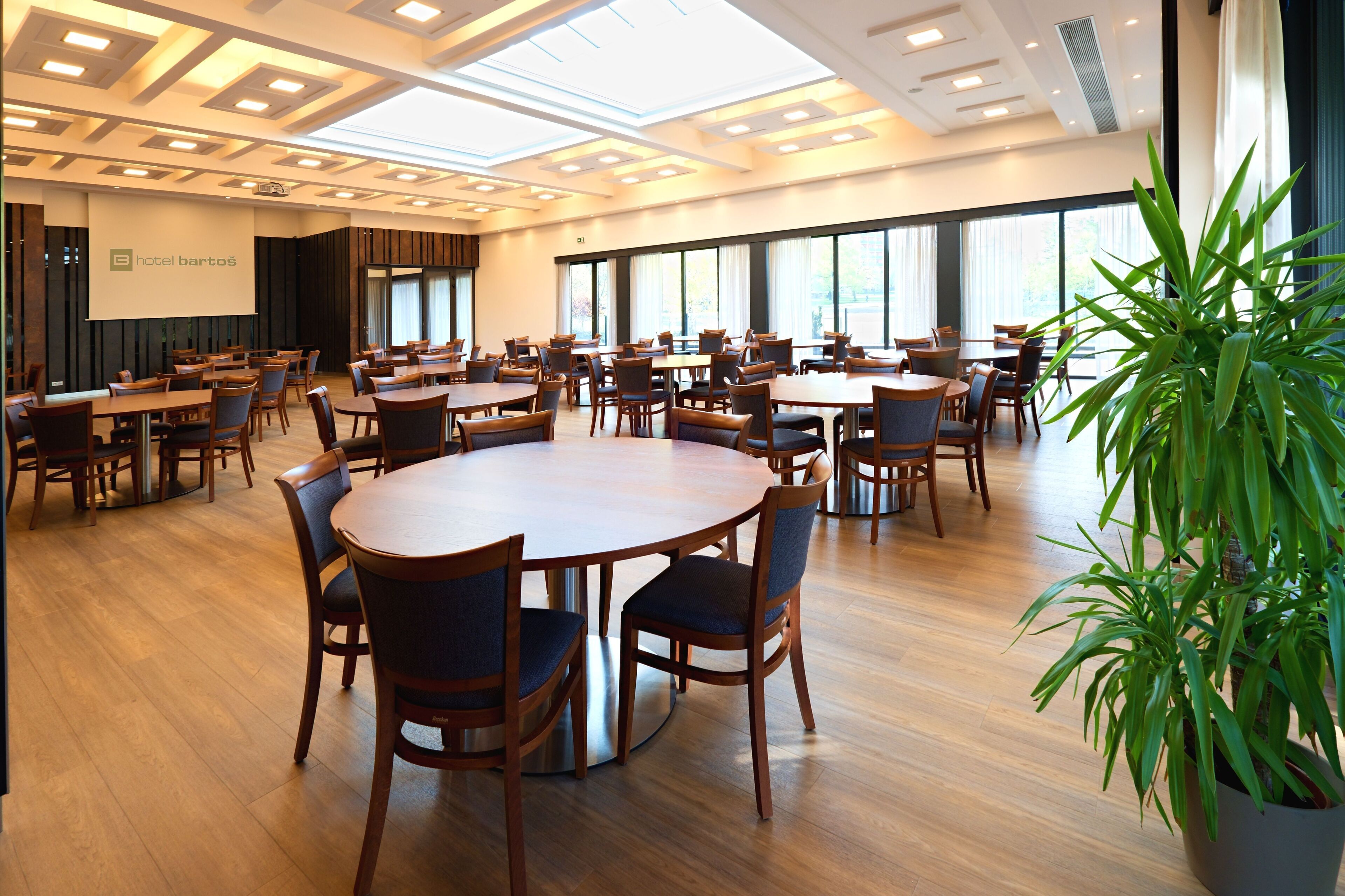 Een ruime vergaderzaal met meerdere tafels en stoelen, grote ramen en een projectiescherm.
