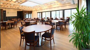 Een ruime vergaderzaal met meerdere tafels en stoelen, grote ramen en een projectiescherm.
