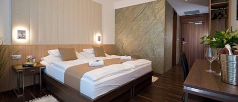 A modern hotel room with a large bed, bedside tables, a desk with a chair, a potted plant, and a wooden wardrobe.