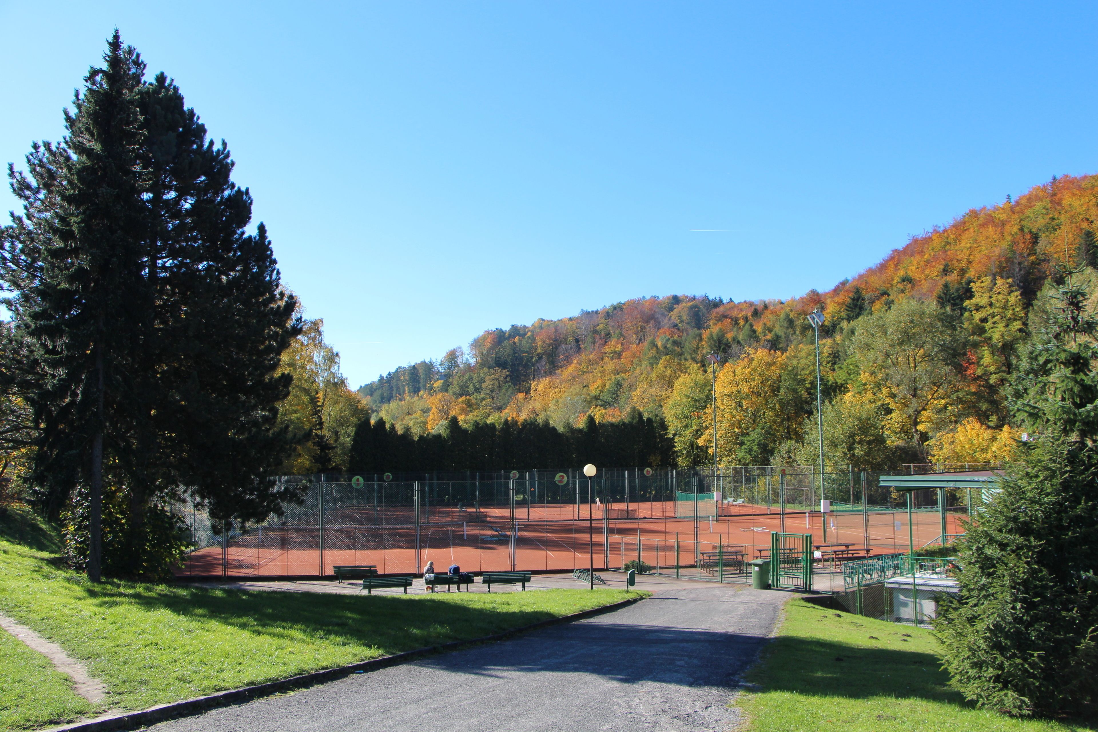 A tennis court with benches and a pathway leading to it, surrounded by trees and a hill.