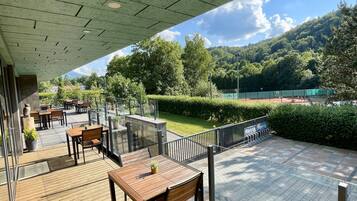 An outdoor seating area with wooden tables and chairs, a glass railing, and a view of a green landscape with trees and a hill.