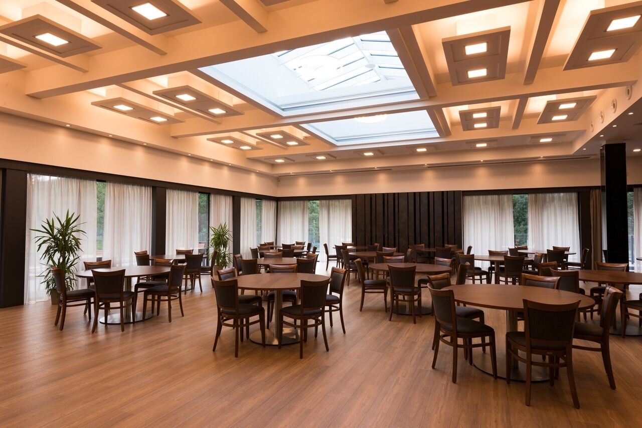 A spacious dining area with multiple tables and chairs, large windows with curtains, and a skylight.