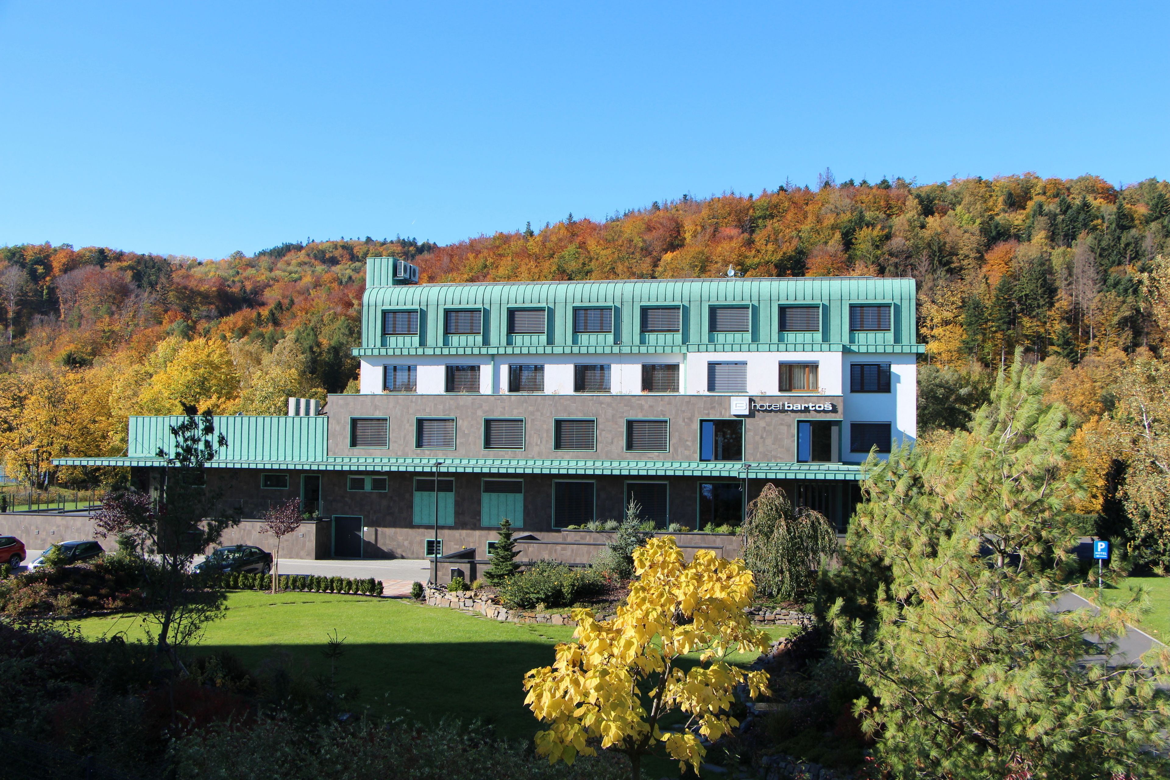 Hotel de vários andares com telhado verde e paredes brancas, rodeado por folhagem de outono.