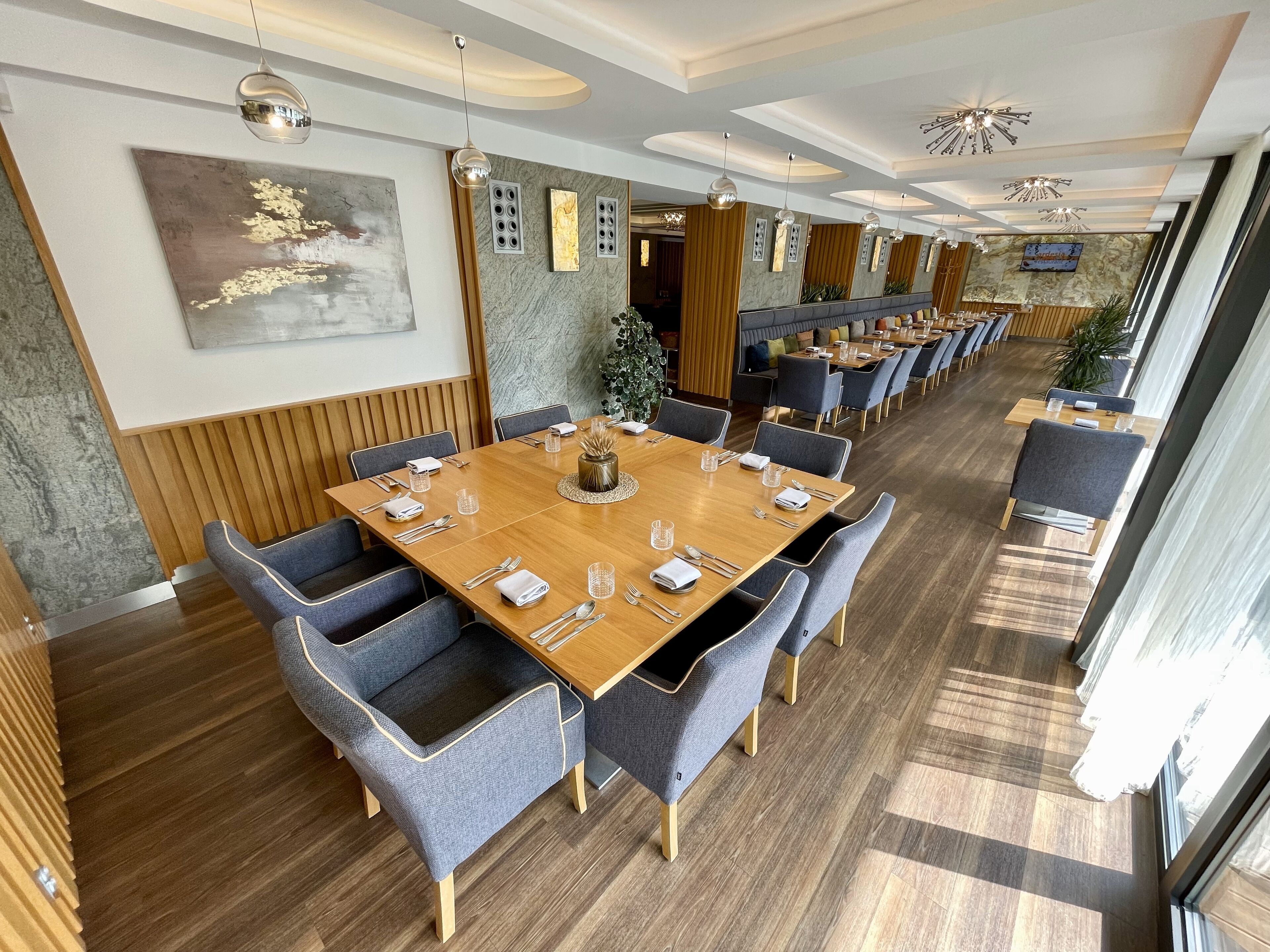 A spacious dining area with long wooden tables, grey upholstered chairs, and modern pendant lighting.