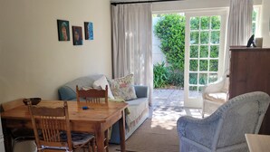Un salón acogedor con una mesa de comedor de madera, un sofá y una silla de mimbre. Hay un ventanal con cortinas, una puerta que da al exterior y un mueble pequeño.