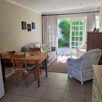 Un salón acogedor con una mesa de comedor de madera, un sofá y una silla de mimbre. Hay un ventanal con cortinas, una puerta que da al exterior y un mueble pequeño.