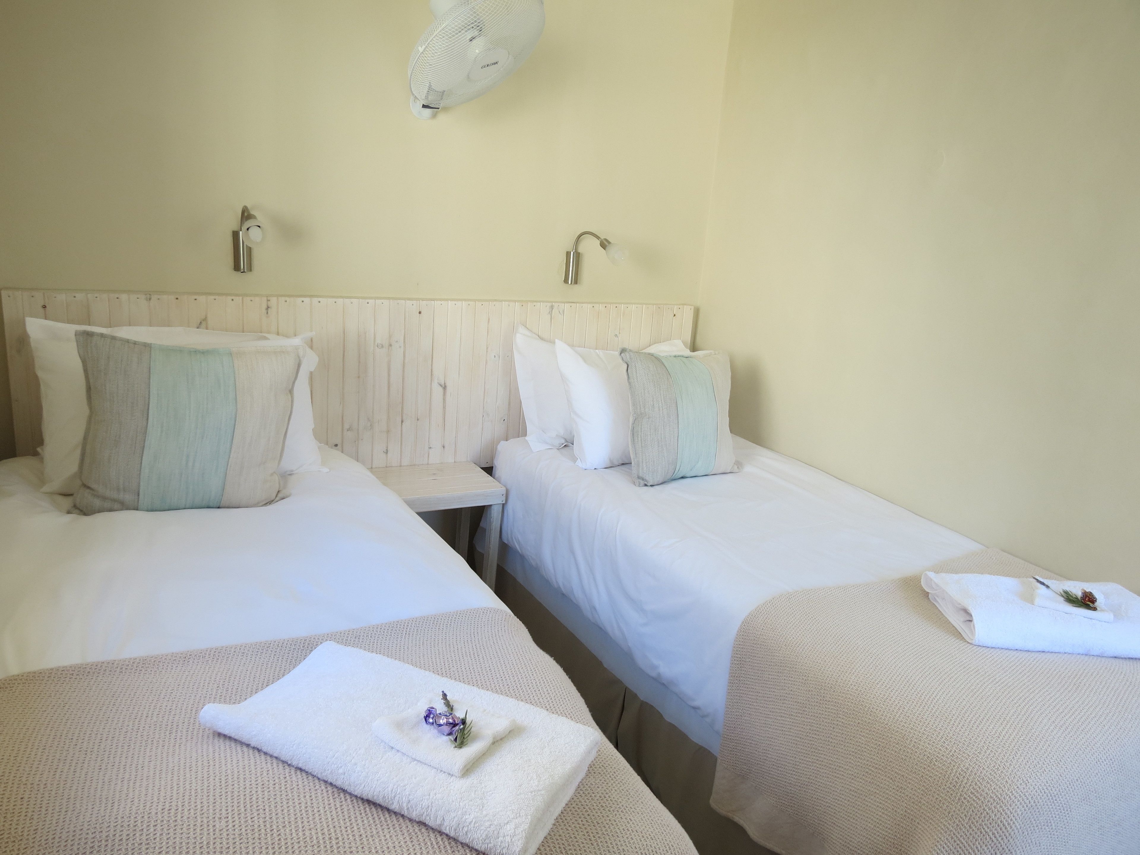A hotel room with two beds, each with white bedding and a wooden headboard. There is a small table between the beds, and a ceiling fan above.