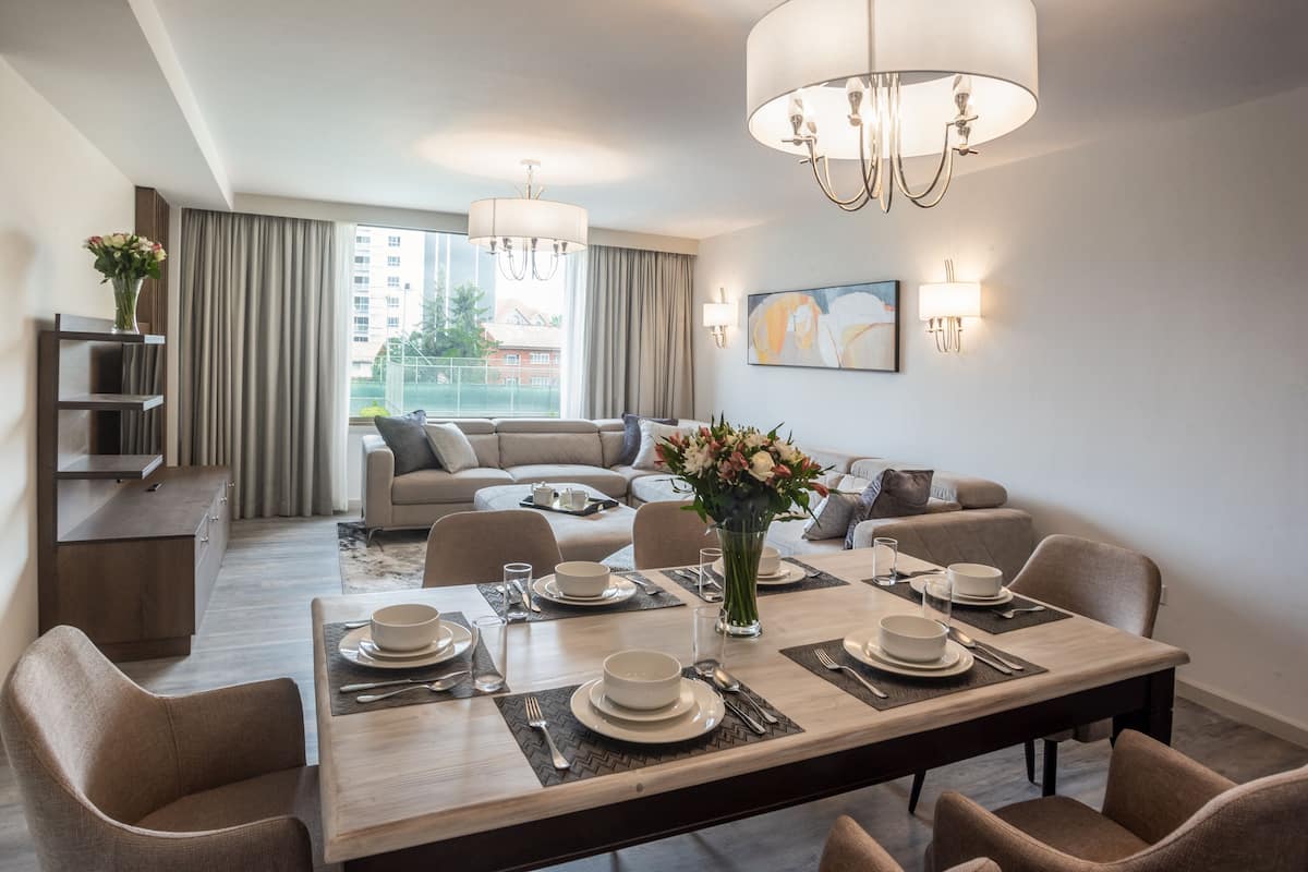A modern hotel room with a dining area, a sofa set, a large window with curtains, and a chandelier.
