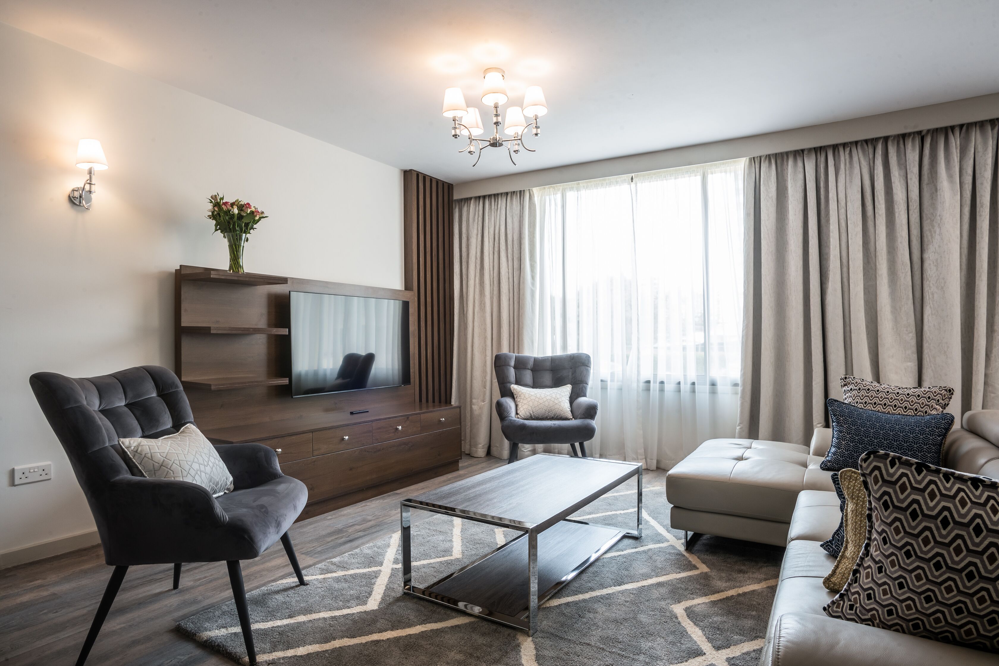 A modern living room with a sofa, two armchairs, a coffee table, a TV on a wooden cabinet, and a chandelier.