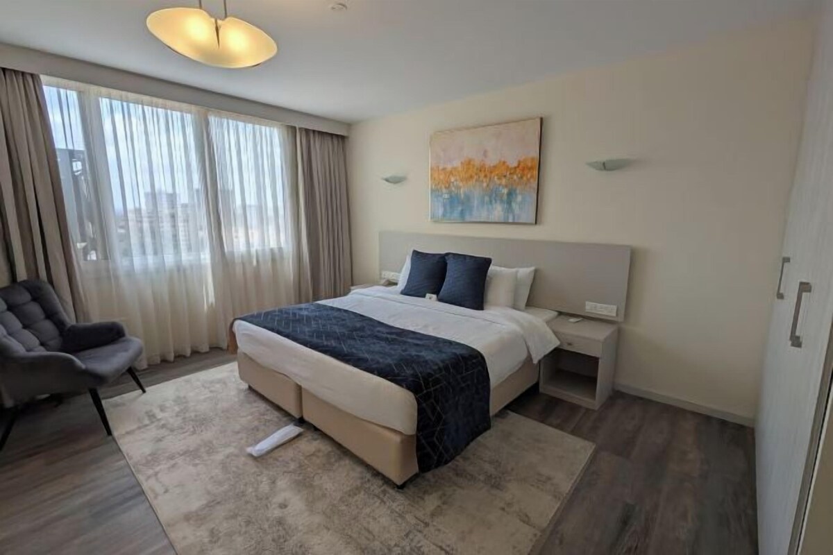 Quarto de hotel com uma cama grande, uma cadeira, uma escrivaninha e um quadro na parede.