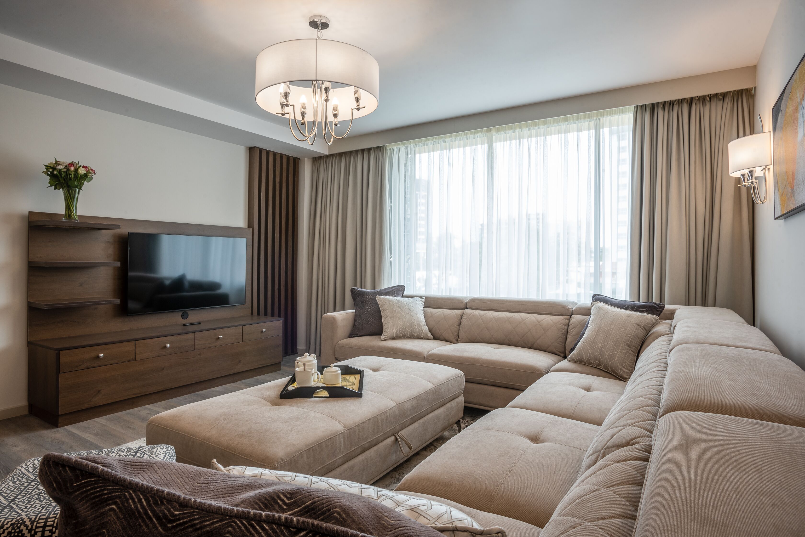 A modern living room with a large sectional sofa, a flat-screen TV, a wooden entertainment unit, and a chandelier.
