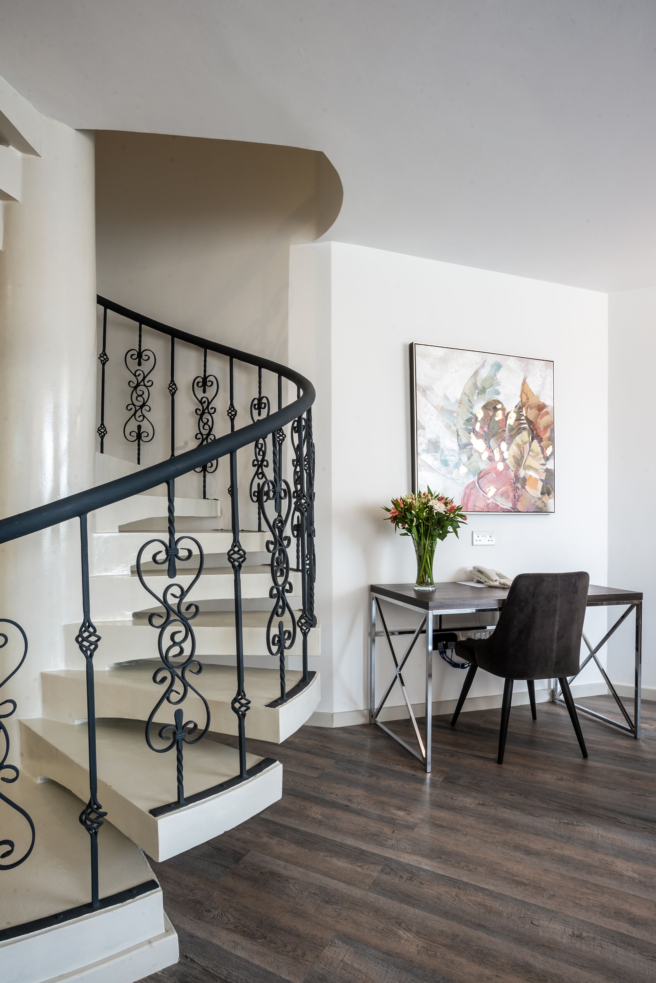 Una escalera con barandillas negras ornamentadas, un escritorio con un florero y una obra de arte enmarcada en la pared.