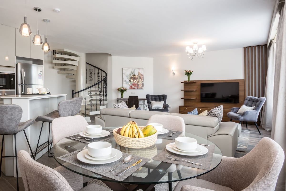 A modern living room with a glass dining table, grey sofas, and a kitchen with a built-in oven and microwave.