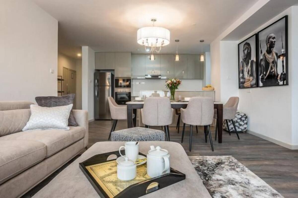 A modern living room with a sofa, a coffee table with a tray, a dining area with chairs, and a kitchen in the background.