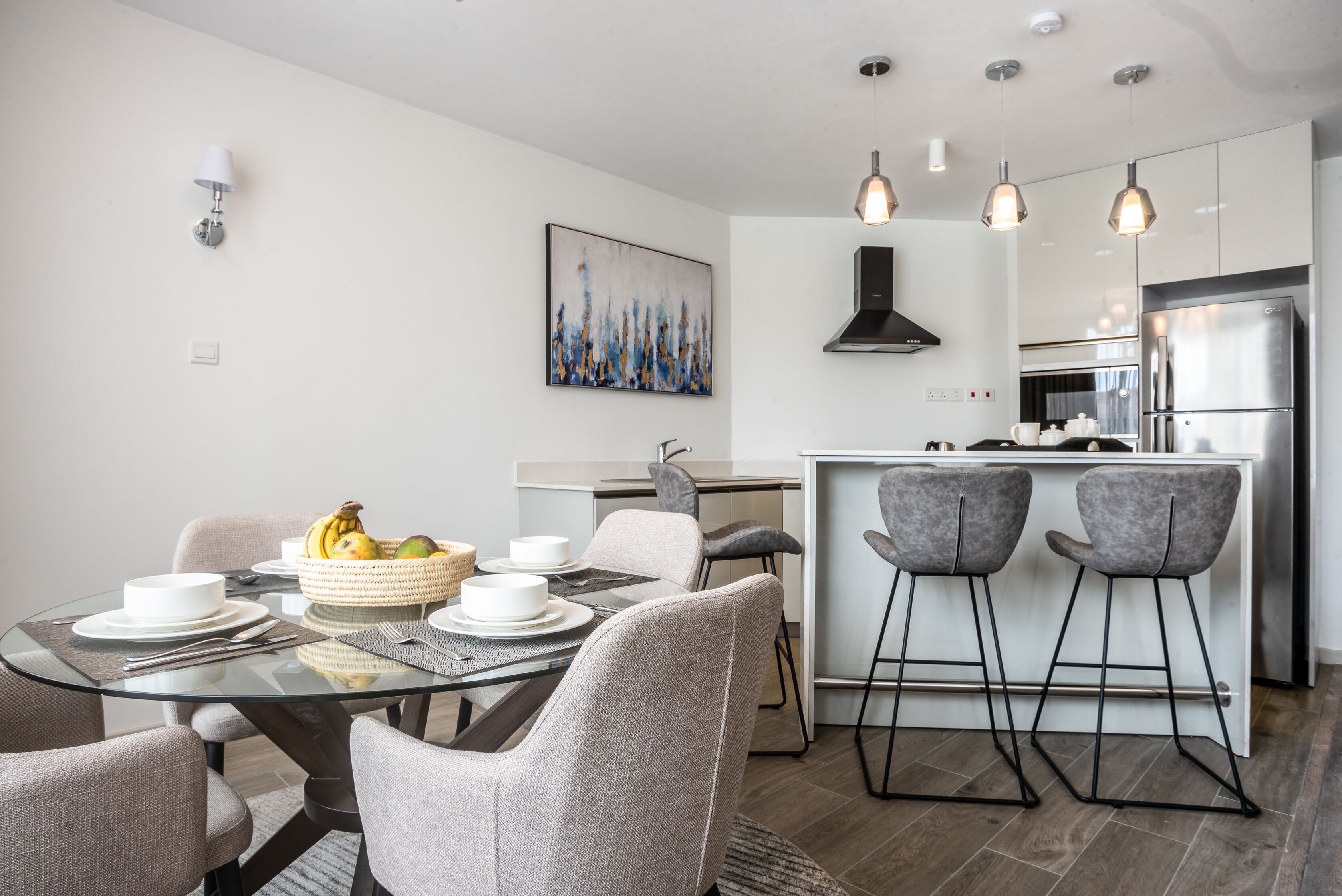A modern kitchen with a stainless steel refrigerator, a dining area with a glass table, and grey upholstered chairs.