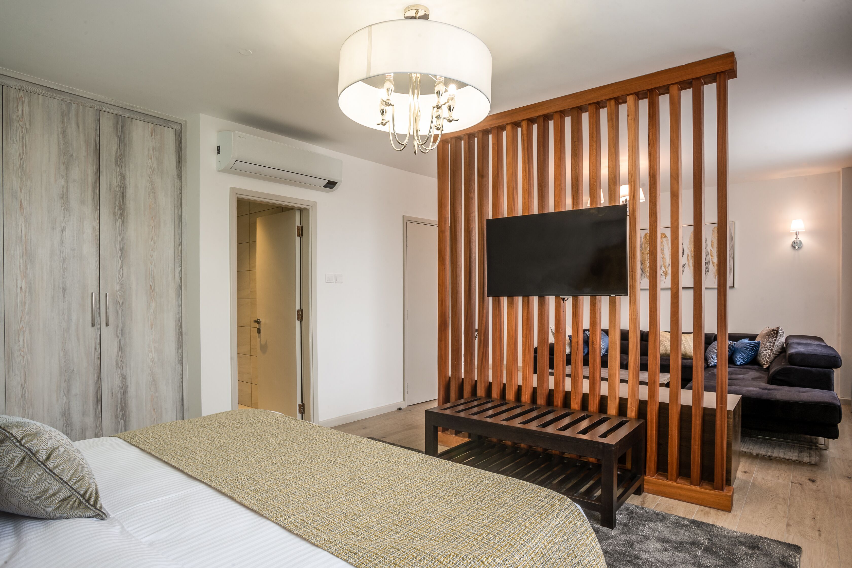 Une chambre à coucher comprenant un lit, un téléviseur fixé sur un panneau en bois, une armoire et un canapé dans le salon attenant.
