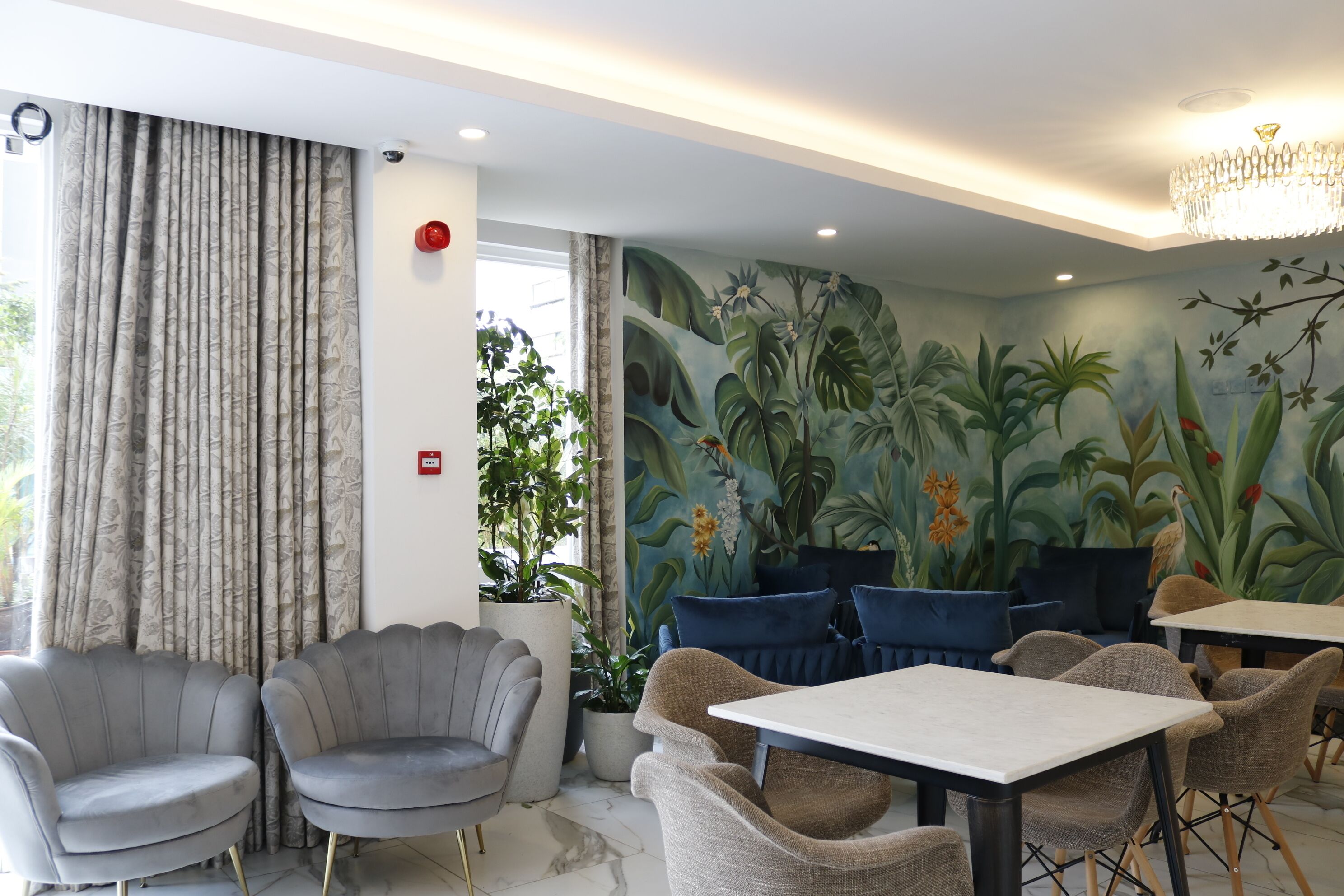 Een modern interieur met een muurschildering van tropische planten, gestoffeerde grijze stoelen, een witte tafel en een kroonluchter.
