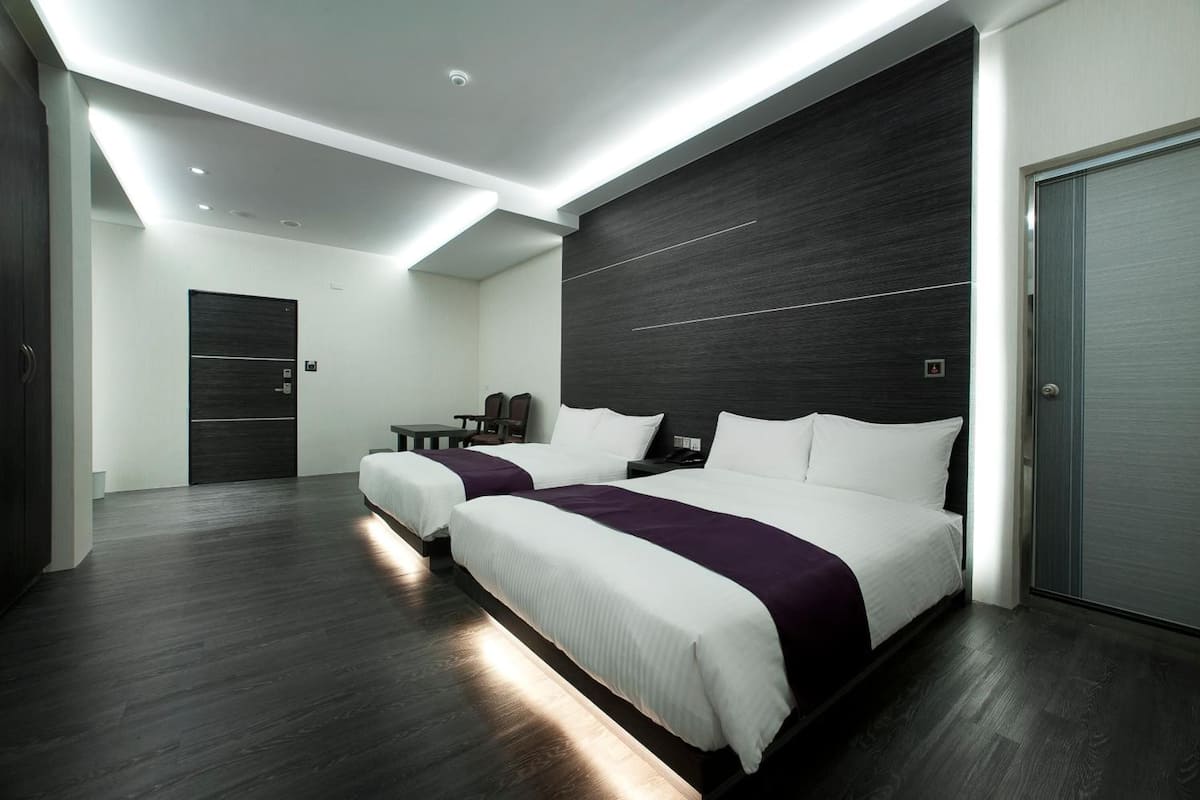 Quarto de hotel moderno com duas camas, piso de madeira escura e uma janela grande.