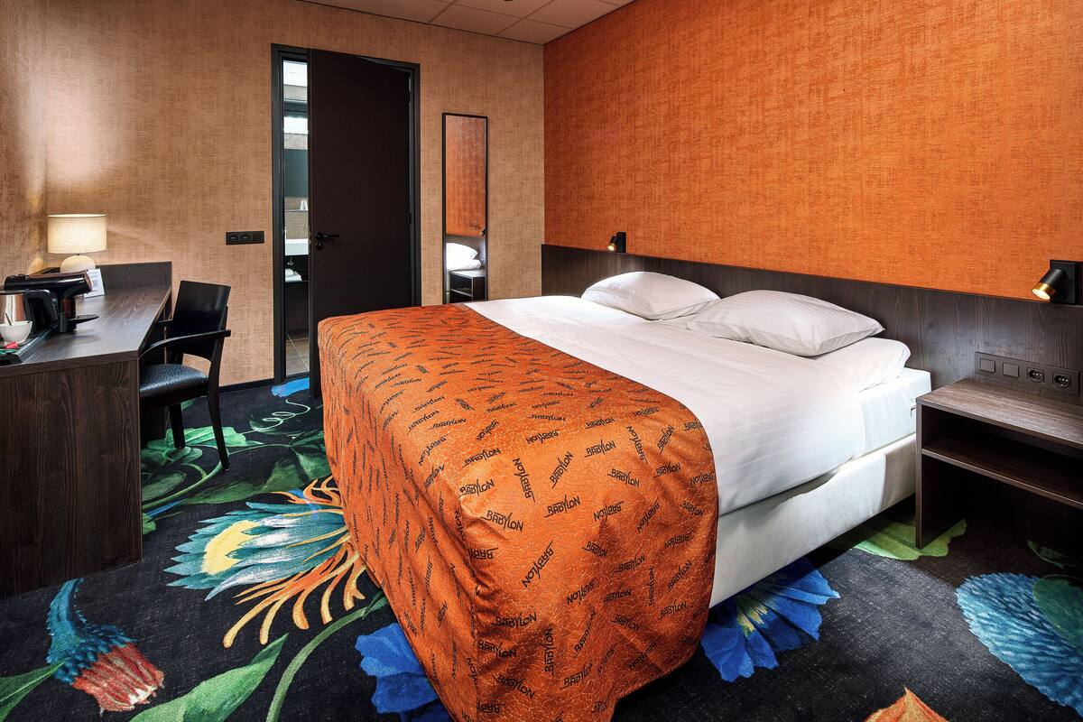 A hotel room with a bed, a desk with a lamp, a chair, and a mirror.
