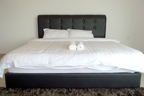 A neatly made bed with white linens and two rolled towels, a dark headboard, and a fluffy rug at the foot of the bed.
