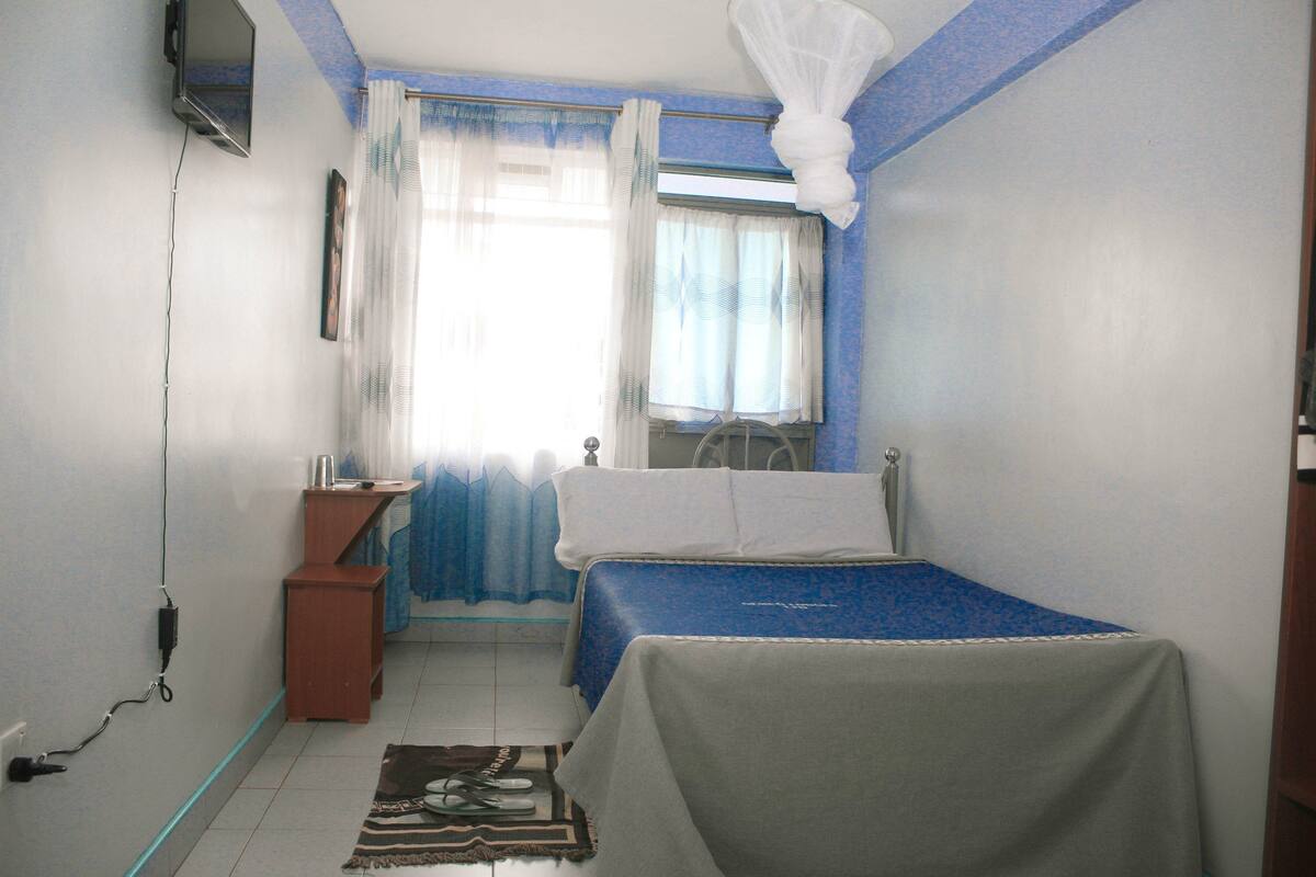 Une chambre avec un lit simple, une table de chevet, une commode, une fenêtre avec des rideaux et un téléviseur fixé au mur.