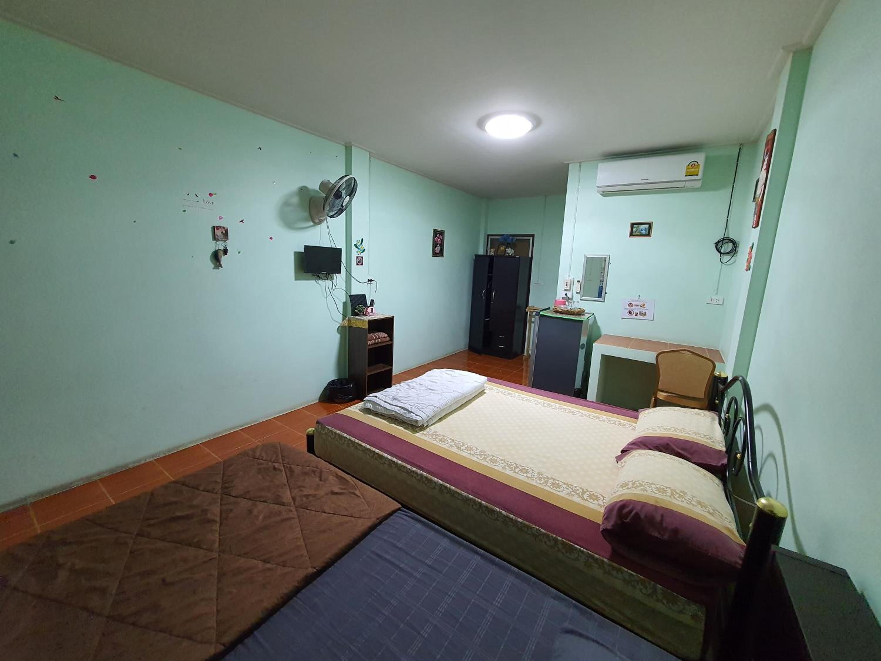 A small, single-room accommodation with a bed, a desk, a wardrobe, and a fan.