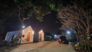 A small, single-story building with a sloped roof, a circular window, and a door. There is a car parked in front, and the area is surrounded by trees and vegetation.