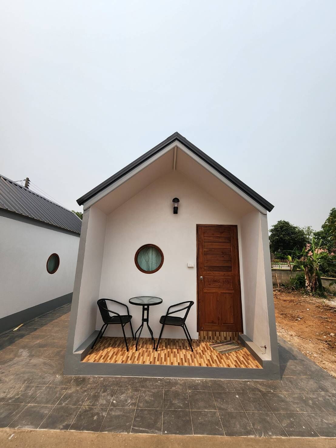 A small, single-story building with a sloped roof, a wooden door, and a small round window. There are two black chairs and a small round table in front of the building.