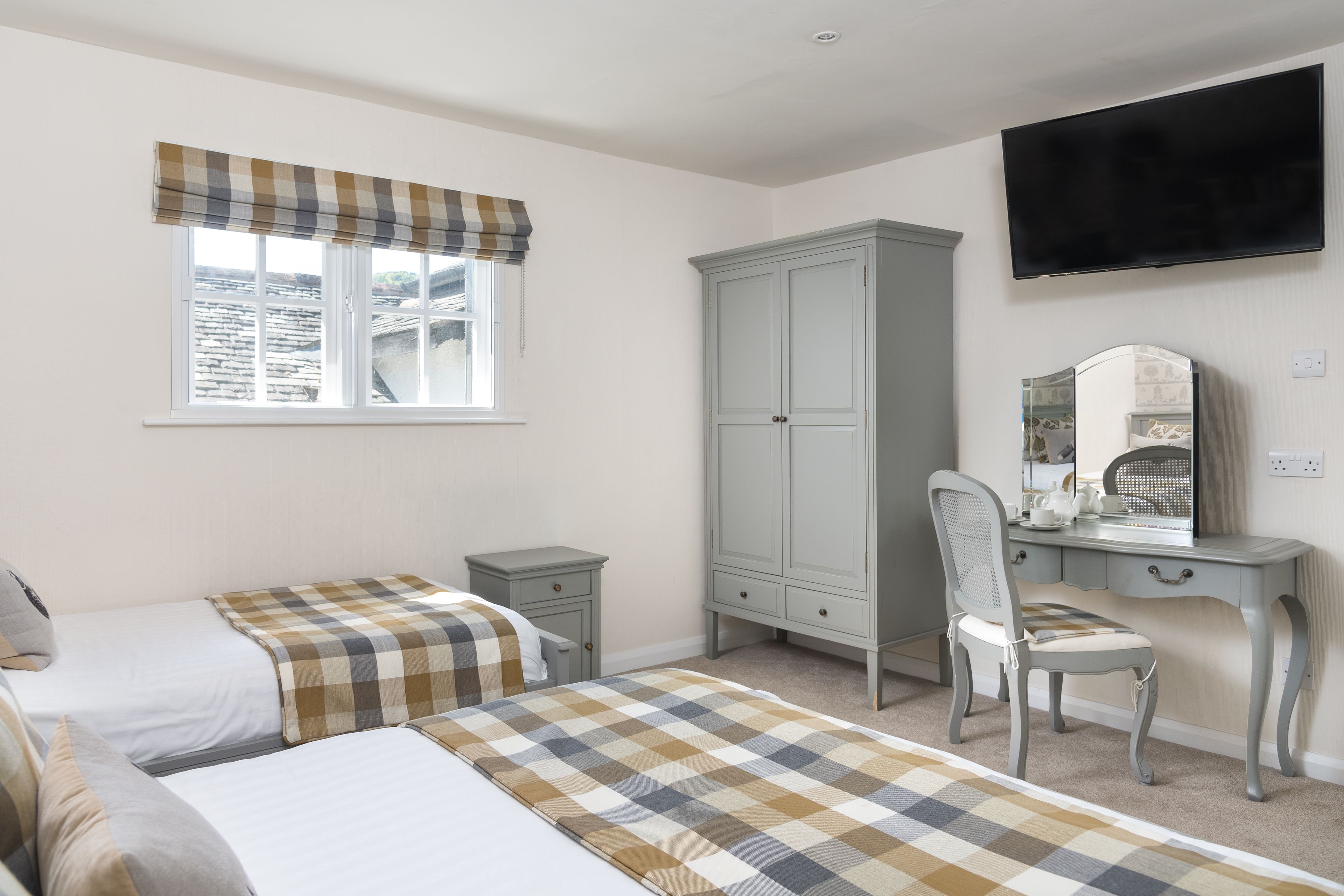 A bedroom with two beds, a wardrobe, a desk with a chair, a television, and a window with a checkered curtain.