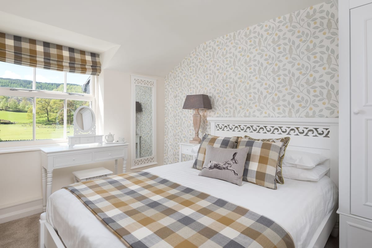 A bedroom with a large bed, a dresser, a mirror, and a window with a view of a green landscape.