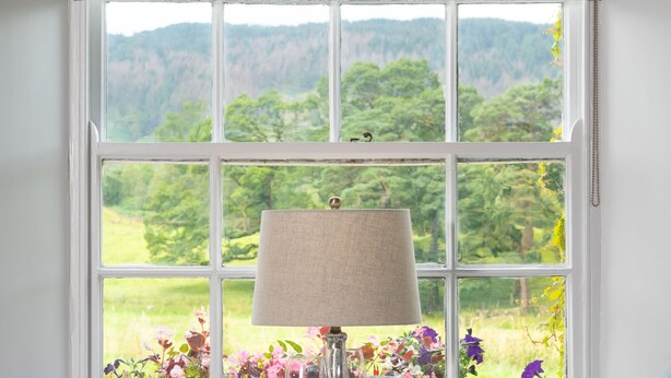 A dining area with a table set for two, a window with a view of greenery, and a decorative window valance.