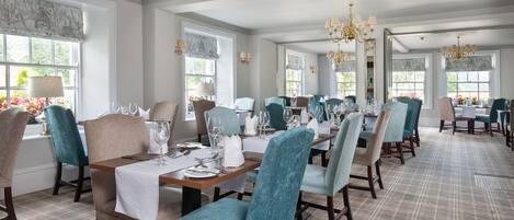 A dining area with multiple tables set for service, chairs upholstered in teal and beige, and chandeliers hanging from the ceiling.