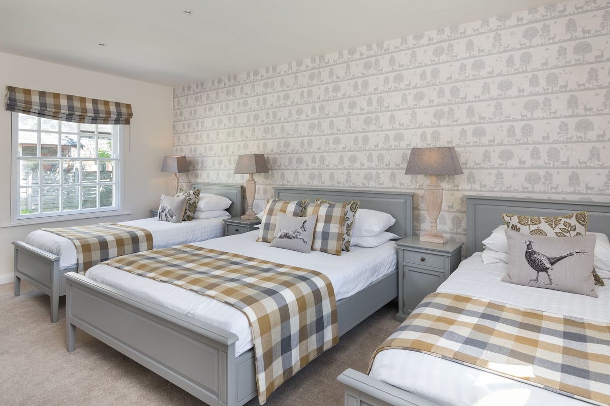 A bedroom with two beds, a window with a checkered valance, patterned wallpaper, and bedside lamps.