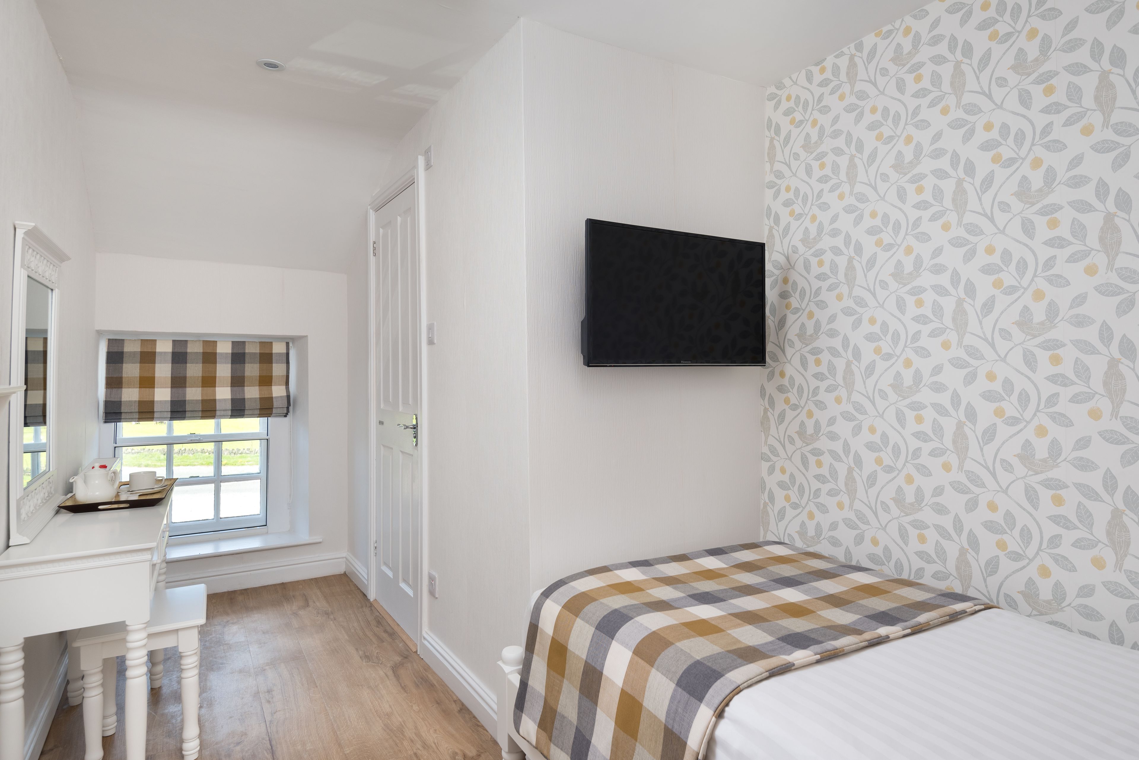 Un dormitorio con cama, televisor montado en la pared, ventana con persiana a cuadros y escritorio blanco con silla.