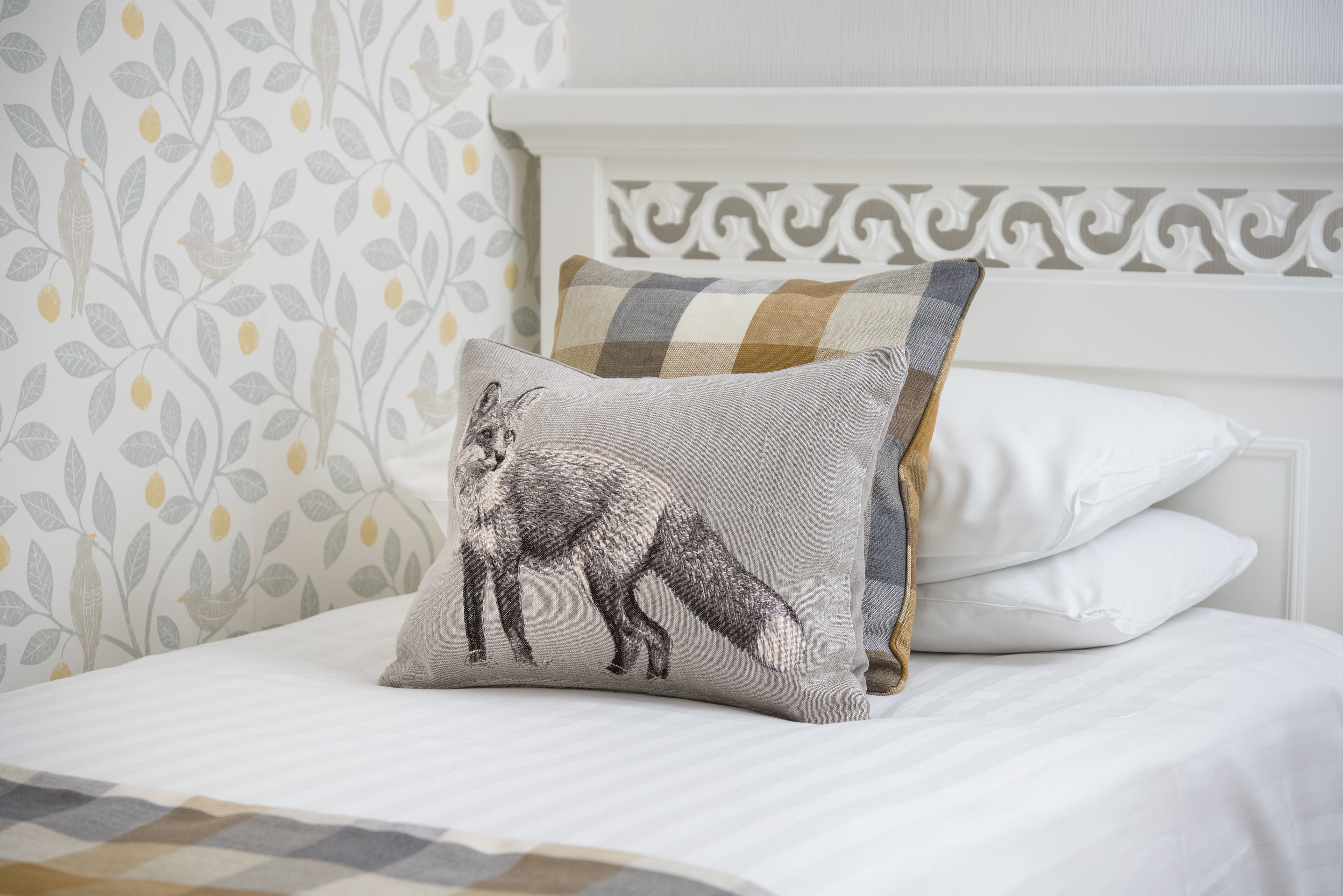 A bed with a white headboard, a pillow with a fox design, and a patterned wallpaper.