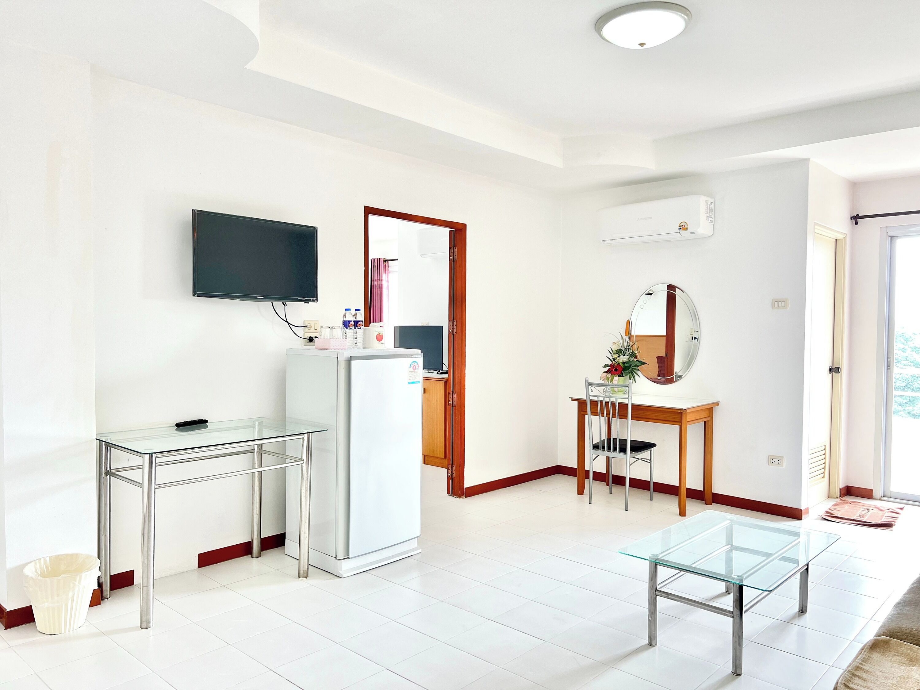 Une chambre d’hôtel moderne avec une table en verre, un réfrigérateur, un téléviseur fixé au mur et une chaise en bois.