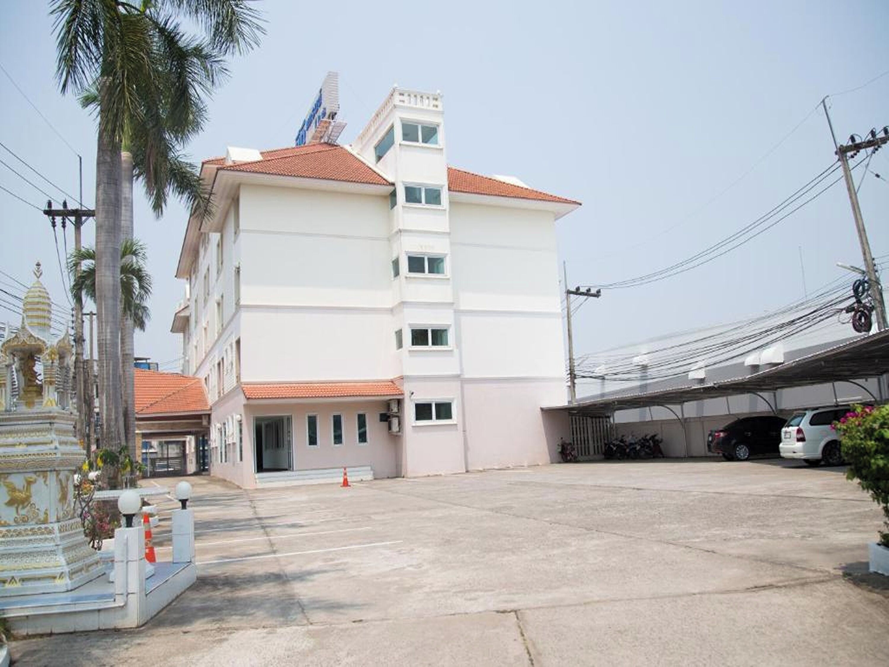 A multi-story building with a flat roof, a parking area, and a small shrine-like structure to the left.