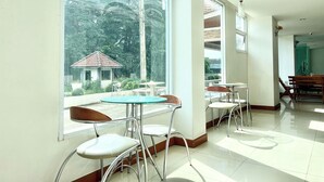 A bright, sunlit room with large windows overlooking a lush garden and a small building. There are several tables and chairs arranged for dining or socializing. - BT Grand Condotel (Udon Thani)